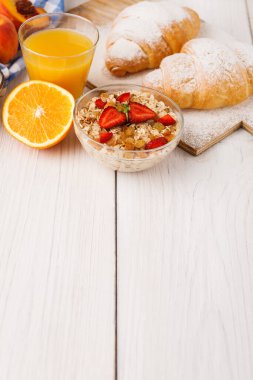 Kolaj, kruvasan, bir bardak meyve suyu, bir kase tahıl ve ahşap bir masada taze meyve içeren kahvaltı setini gösterir. Sahne basit ve davetkar..