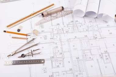 Detaylı mimari planların üzerinde bir dizi taslak araç yatıyor. Ögeler arasında kalemler, bir cetvel, bir pusula, silgi ve rulo kağıtlar var. Çalışma alanı tasarım görevleri için düzenlenmiştir.