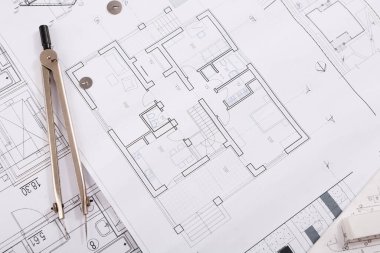 Bir planlar koleksiyonu ve mimari planlar bir masanın üzerine yayılır. Pusula ve çizim malzemeleri gibi araçlar görülebilir. Çalışma alanı meşgul ve tasarıma odaklanmış görünüyor.