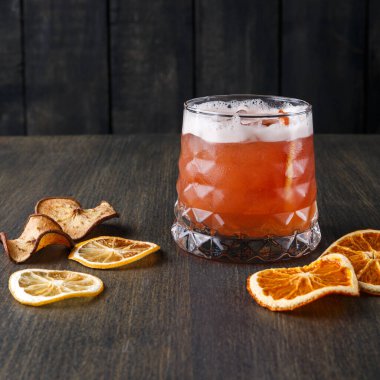 Yanında kurumuş portakal ve elma dilimleri olan ahşap bir masada bir kokteyl duruyor. İçki zengin bir renk ve üzerinde köpük var. Akşam toplantıları için sıcak bir ortam sunuyor..