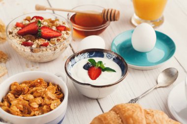 Farklı yiyecek ve içecekler ahşap bir masada sergilenir. Sahnede yoğurt, meyve, granola, yumurta, bal ve meyve suyu yer alıyor. Bu yemek için tipik bir kahvaltı düzenlemesi..
