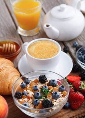 Kahvaltı malzemeleri ahşap bir masanın üzerinde düzenlenir. Bir kase meyve ve tahıl, bir fincan kahve, bir bardak meyve suyu ve bir kruvasan var. Sahne, sabah için yemeği gösteriyor..