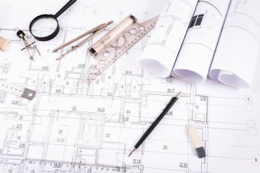 Ayrıntılı taslak araçları olan bir masanın üzerine planlar konur. Kalemler, pusulalar ve cetveller görünür, tasarım çalışmalarına hazırlık gösteriyor. Planlar ayrıntılı olarak görülebilir..