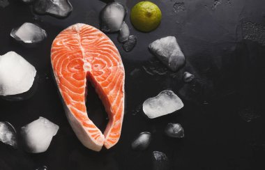 Bir somon kesiği, buzla ve limonla çevrili karanlık bir yüzeye yerleştirilir. Sahne yiyeceklerin dokularını ve renklerini ön plana çıkarıyor. Bu kurulum taze malzemelerin bir koleksiyonunu gösterir.