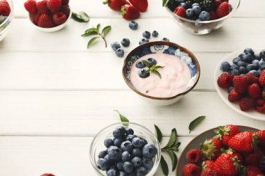 Bir kase seti çilek, ahududu, böğürtlen ve yabanmersini gibi çeşitli taze meyveler içerir. Yaban mersinli yoğurt beyaz bir masanın ortasında duruyor..