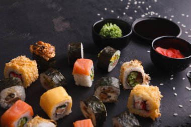 Suşi ruloları çeşitli malzeme ve malzemelerle karanlık bir yüzeye yerleştirilir. Suşiye küçük kaseler wasabi, soya sosu ve turşulu zencefil eşlik ediyor. Yemek için ortam hazır görünüyor..