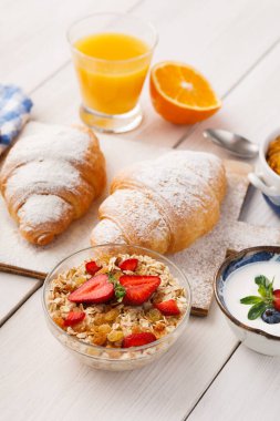 Kahvaltıda iki kruvasan, bir kase çilekli granola, yoğurt ve masada portakal suyu var. Ayar, doğal ışıkla sıcak bir his veriyor..