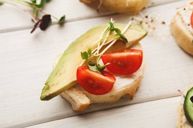 Dilimlenmiş avokado, kiraz domatesleri ve lahanalarla kaplı çeşitli küçük ekmek parçaları açık ahşap bir yüzeye kurulmuştur. Bu koleksiyonda basit ve sağlıklı atıştırmalıklar sergileniyor..