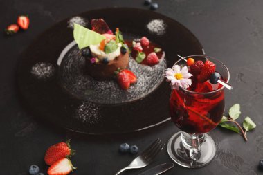 Karanlık bir yüzeye yerleştirilmiş bir tatlı ve içecek setinde meyveli bir çikolatalı tatlı ve dutlu bir içecek yer alır. Basit süslemeler ekrana renk ekler.