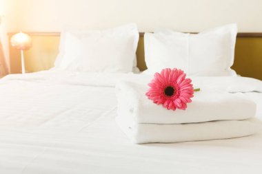 Beyaz havlu koleksiyonu düzgünce yatağa yerleştirildi. Pembe bir çiçek havluların üzerinde oturur, biraz renk katar. Odanın yumuşak ışıklandırması ve rahat bir atmosferi var..