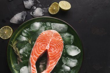 Bir somon bifteği buz küpleriyle dolu yeşil bir tabağa yerleştirilir. Tabakta limon dilimleri ve taze bitkiler de var. Ayar karanlık bir yüzeye sahip..