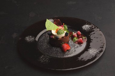 Bir tatlı karanlık bir tabakta oturur. Tatlıda çikolata, krema ve yeşil süsleme var. Taze böğürtlen ve nane şekeri tatlının etrafını sarar. Bu yemeğin güzel bir yemek için cazip bir tasarımı var..