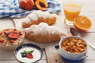 Sahne, tahtadan bir masada kruvasan, meyve, yoğurt ve mısır gevreği içeren kahvaltı malzemeleri koleksiyonunu gösteriyor. Bir bardak meyve suyu yemeğin yanında oturur ve bir sabah havası yaratır..