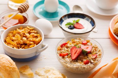 Farklı kahvaltı öğeleri masanın üzerine serilmiştir. Mısır gevreği, meyveli yoğurt, yumurta, bal ve taze ekmek var. Bu ortam sabah yemekleri ve atıştırmalıklar içindir..