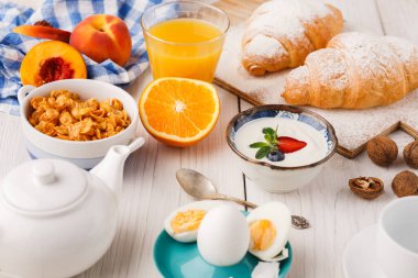 Kahvaltı malzemelerinin bir koleksiyonu ahşap bir masada düzenlenir. Kruvasan, taze meyve, yoğurt, yumurta, mısır gevreği ve meyve suyu birlikte sergileniyor ve çeşitli sabah yiyecekleri sergileniyor..