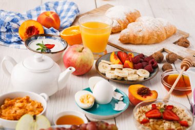 Sahne bir masaya yerleştirilmiş çeşitli kahvaltı öğelerini gösteriyor. Meyveler, pastalar, içecekler ve biraz mısır gevreği var. Düzenleme renkli ve davetkar..