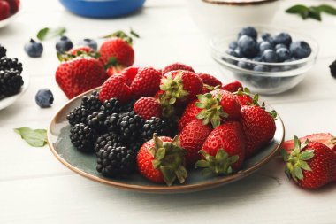 A set of fresh strawberries and blackberries sits on a plate. Blueberries are in small bowls around the plate. The scene shows vibrant berries on a simple table.