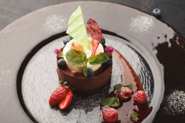 Çikolatalı bir tatlı siyah bir tabağa konur. Sergide taze böğürtlen ve nane ile süslenmiş kek ve krema katmanları yer alıyor. Burası bir restoran, davetkar bir ortam yaratıyor..