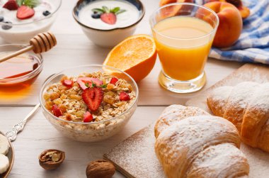 Masaya yerleştirilmiş kahvaltı malzemeleri koleksiyonu. İçinde yoğurt, meyve, granola, portakal suyu ve taze hamur işleri olan kaseler var. Sabah oldu ve yemekler davetkar görünüyor..