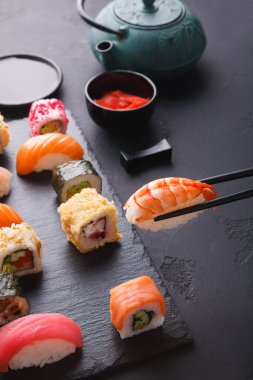 Suşi parçaları bir tabağa dizilir. Farklı suşi türleri sergilenir ve karides yemek çubukları kullanılarak alınır. Yakınlarda bir çaydanlık ve sos var..