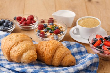 Kahvaltı malzemelerinin bir koleksiyonu masanın üzerinde kurulur. Kruvasanlar kareli bir kumaşın üzerine konur. Kaselerde çeşitli meyveler, yoğurt ve tahıl gevreği var. Kahve beyaz bir fincanda servis edilir..