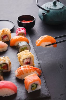 Siyah bir tahta üzerine hazırlanmış bir suşi seti. Ekmek ve sashimi dahil olmak üzere farklı suşi türleri mevcuttur. Suşinin yanında yeşil bir çaydanlık ve yanında küçük bir kase sos var..