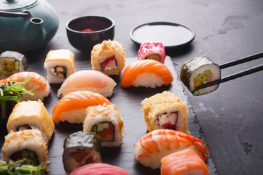 Siyah bir tahta yüzeyde çeşitli suşi türleri sergilenir. Balık, pirinç ve sebzelerden yapılmış rulo ve parçalar var. Çay ve soslar yakınlarda, yemek hazırlıyorlar..