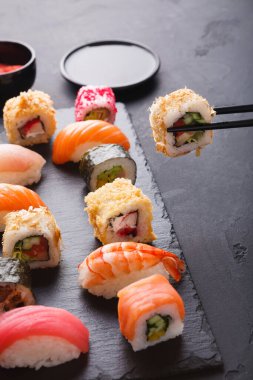 Bir suşi seti siyah bir levhada durur. Bir koleksiyonda farklı suşi türleri düzenlenir. Renkli rulo ve parçalar var. Bir rulo çubukla tutulur. Yakınlarda küçük bir kase var..