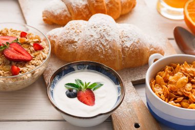 Kolaj, üzerinde kruvasan, meyveli yoğurt ve mısır gevreği olan kahvaltı setini gösteriyor. Masanın sıcak bir güneş ışığı görünüşü var. Sahne neşeli bir sabah yemeğini vurguluyor..