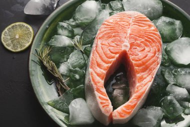 Dekor yeşil bir tabakta buzun üzerine yerleştirilmiş somon filetosu koleksiyonunu gösteriyor. Pişirmeye hazırlanan taze limon dilimleri ve otlar balığa eşlik eder. Olay yeri canlı bir yemek düzeneğini vurguluyor..