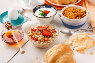 Kahvaltı malzemeleri bir masanın üzerinde düzenlenir. Kaselerde yulaf, yoğurt ve mısır gevreği var. Yakında yumurta, bal ve kruvasan var. Taze meyveler de mevcuttur.