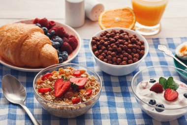 Kahvaltı malzemelerinin bir koleksiyonu bir masanın üzerinde düzenlenir. Sahnede kruvasan, meyve, yoğurt, mısır gevreği ve meyve suyu yer alıyor. Bu kurulum çeşitli sabah yemekleri seçenekleri sunuyor..