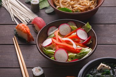 Bir kasede taze sebze ve suşi var. Yakınlarda, başka bir kasede erişte var. Çubuklar masanın üzerinde dinleniyor. Bir yemek için çeşitli renkli yiyecekler sunulur.