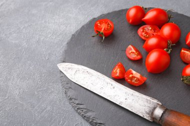 Kırmızı domatesler gri bir kesim tahtasına oturur. Bazı domatesler bütün, bazıları ise küçük parçalara bölünmüş. Yakınlarda yemek pişirmeye hazır bir bıçak duruyor..