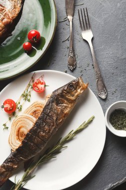 Bir tabak, dilimlenmiş soğanlar, kiraz domatesleri ve taze otların yanında pişmiş bir balık tutar. Ayarlar arasında çatal bıçak ve arka planda yeşil bir tabak yer alıyor.