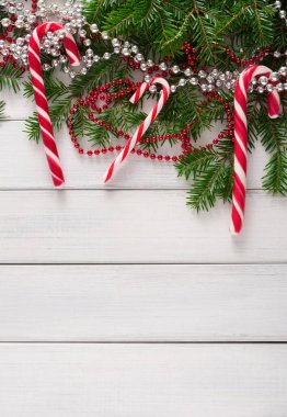 Çam dalları ve parlak boncuklar arasında beyaz ahşap bir yüzeye üç kırmızı ve beyaz baston şeker uzanır. Bu sahne Noel kutlamalarının ve Noel dekorasyonunun ruhunu yansıtıyor..