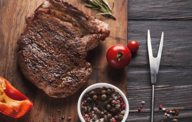 Ahşap bir tahtanın yanında ızgara biftek, baharatlar ve taze sebzeler sergileniyor. Bir kase karışık baharat ve çatal da mevcut, bu da mutfak ortamında yemek hazırlığı olduğunu gösteriyor..