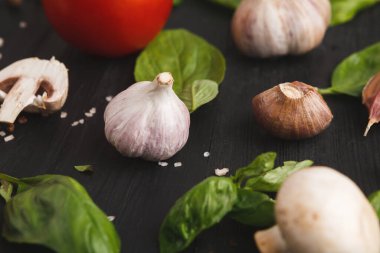 Sebzeler ve bitkiler karanlık bir yüzeye yerleştirilir. Sarımsak, mantar ve domates fesleğen yapraklarıyla çevrilidir. Bu kurulum yemek pişirmeye hazır malzemeleri gösterir.