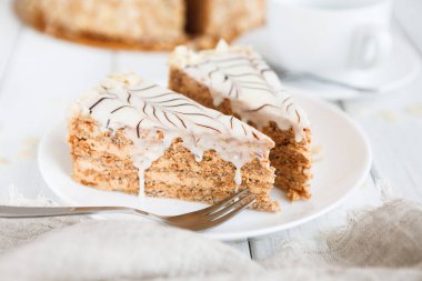Yanında çatalla beyaz bir tabakta oturan iki dilim pasta. Pastanın katmanları zengin dolgu ve tatlı krema gösteriyor. Arka planda rahat bir ortam ve yakınlarda bir fincan görülüyor..