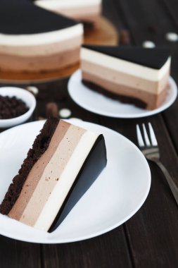 Farklı katmanlı pastalar, beyaz tabakların üzerinde, ahşap bir masadaki tüm pastanın yanında duruyor. Çikolata, krema ve diğer tatlar tatlı severler için renkli bir vitrin oluşturur..