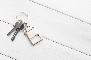 Anahtarlar ev şeklindeki anahtarlık takılı ahşap bir yüzeyde duruyor. Aydınlık ve basit bir ortam. Bu sahne, evlere veya binalara girmek için kullanılan günlük öğeleri gösterir.
