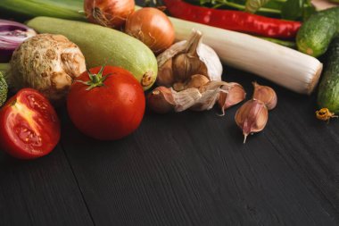 Koyu ahşap bir masanın üzerine çeşitli sebzeler yayılır. Bu karışımda domates, sarımsak, soğan, kabak ve bitkiler yer alıyor. Her sebze farklı şekillerde ve renklidir..