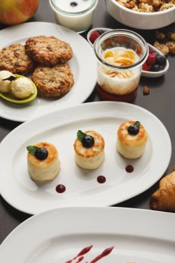 Beyaz tabaklarda çeşitli tatlılar bulunan bir kolaj. Tatlılar arasında mini kek, kurabiye, yoğurt, meyve ve kruvasan yer alıyor. Renkler ve dokular değişik ve iştah açıcı bir görüntü yaratır..