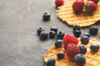 İki waffle karanlık bir yüzeye yerleştirilir. Taze ahududu ve yaban mersiniyle kaplanmışlar. Ayarlar basit ve gıda düzenine odaklı..