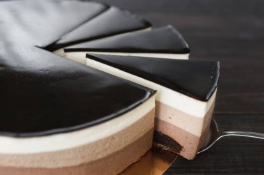 Masanın üzerinde birden fazla çikolata ve krema katmanlı bir pasta duruyor. Bir dilim kesilmiş, farklı katmanları gösteriyor. Etraftaki insanların toplandığı bir tatlı toplantısı olacak..