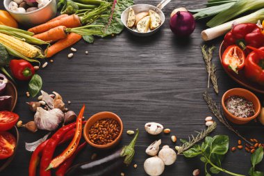 Taze sebzeler, koyu ahşap bir masanın üzerinde dairesel bir düzene yerleştirilir. Havuç, biber, sarımsak ve otlar bir arada karıştırılır. Sahnede yemek pişirmeye hazır malzemeler var..