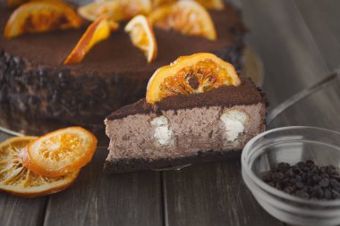 Bir tatlı seti bir dilim kesilmiş bütün bir pastayı içerir. Pastanın çikolata katmanları var ve üzerinde portakal dilimleri var. Tahta bir yüzeyde bir kase çikolata kırıntısı var..