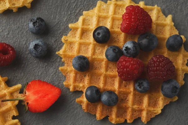 Karanlık bir yüzeye bir waffle seti yerleştirilir. Yaban mersini ve ahududu gibi meyveler waffle 'ların üzerinde düzenlenir. Yakınlarda bir çilek var, renkli bir görüntü oluşturuyor..
