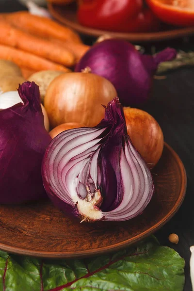 Tahta bir tabakta bir sebze seti düzenlenir. Kırmızı soğan, sarı soğan ve havuç sergilenir. Sahne, taze üretimin doğal dokusunu ve rengini gösteriyor..