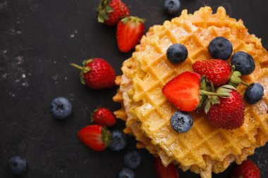 Bir yığın altın waffle karanlık bir yüzeyde duruyor. Taze çilekler ve yaban mersinleri waffle 'ların etrafına saçılırlar. Bu kurgu kahvaltı ya da tatlı için cazip bir sahne yaratır..